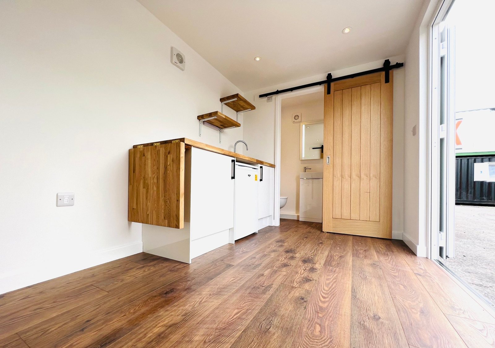 20ft Container Home – One Bed Box, 1 Bedroom – A space-efficient kitchenette with hardwood floors and a sliding barn door leads to the compact bathroom in this 20ft unit