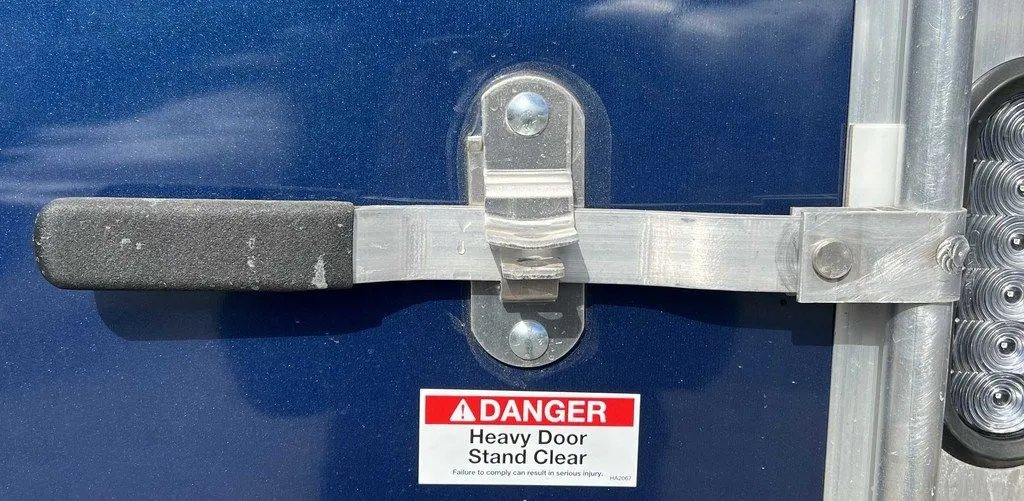 2023 High Country 14 ft Trailer – Blue, Drive-Through – Metal locking bar handle for the ramp door with a safety warning label