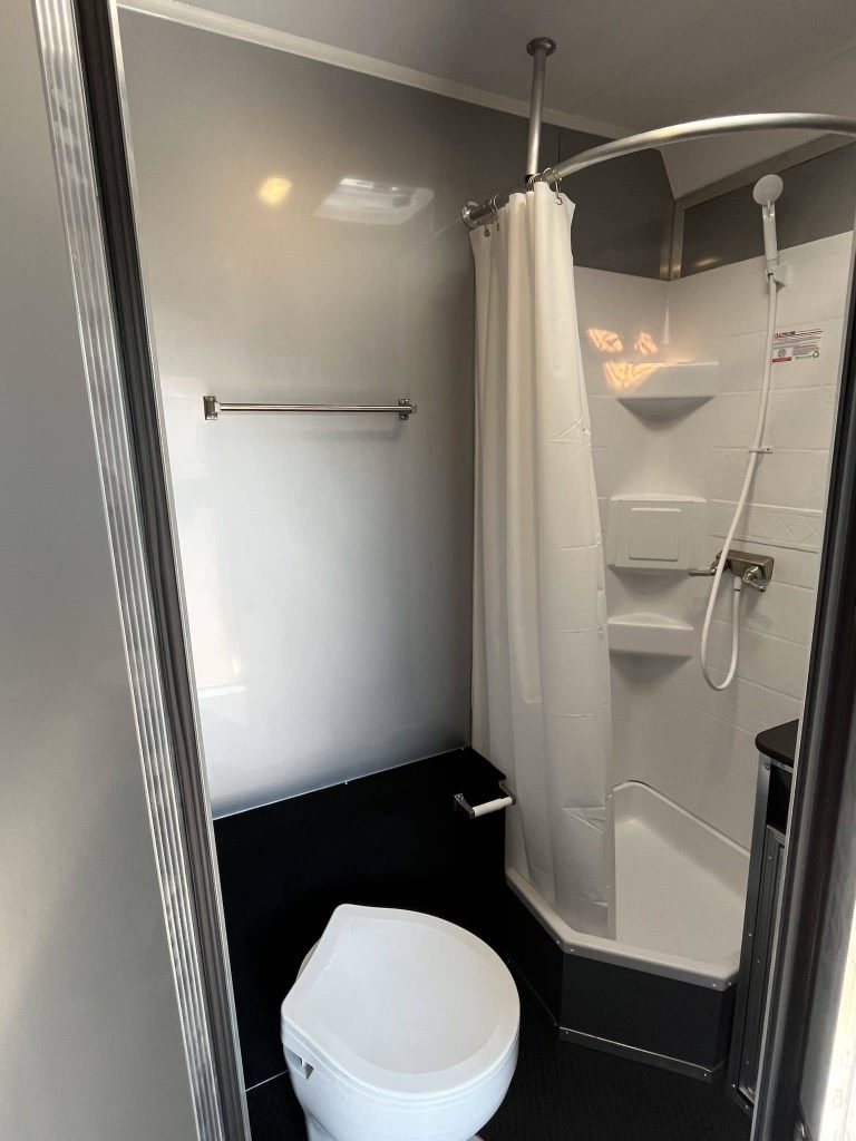 2025 Millennium 34 ft Car Hauler – Corner Bathroom – Interior bathroom with white toilet and corner shower unit with white curtain