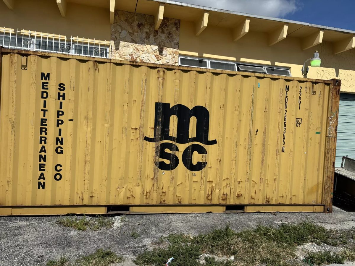 20ft Used Shipping Container – Yellow/Gold, Double Cargo Doors – Weathered yellow shipping container side with dents and rust beside a building