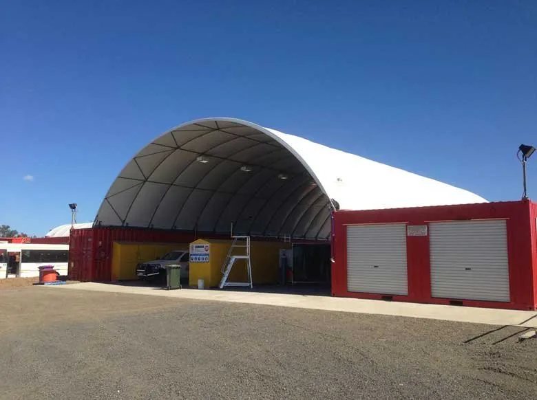 Container Shelter – Domed Tensile Membrane Cover – Large white arched container shelter spanning between two red shipping containers on gravel