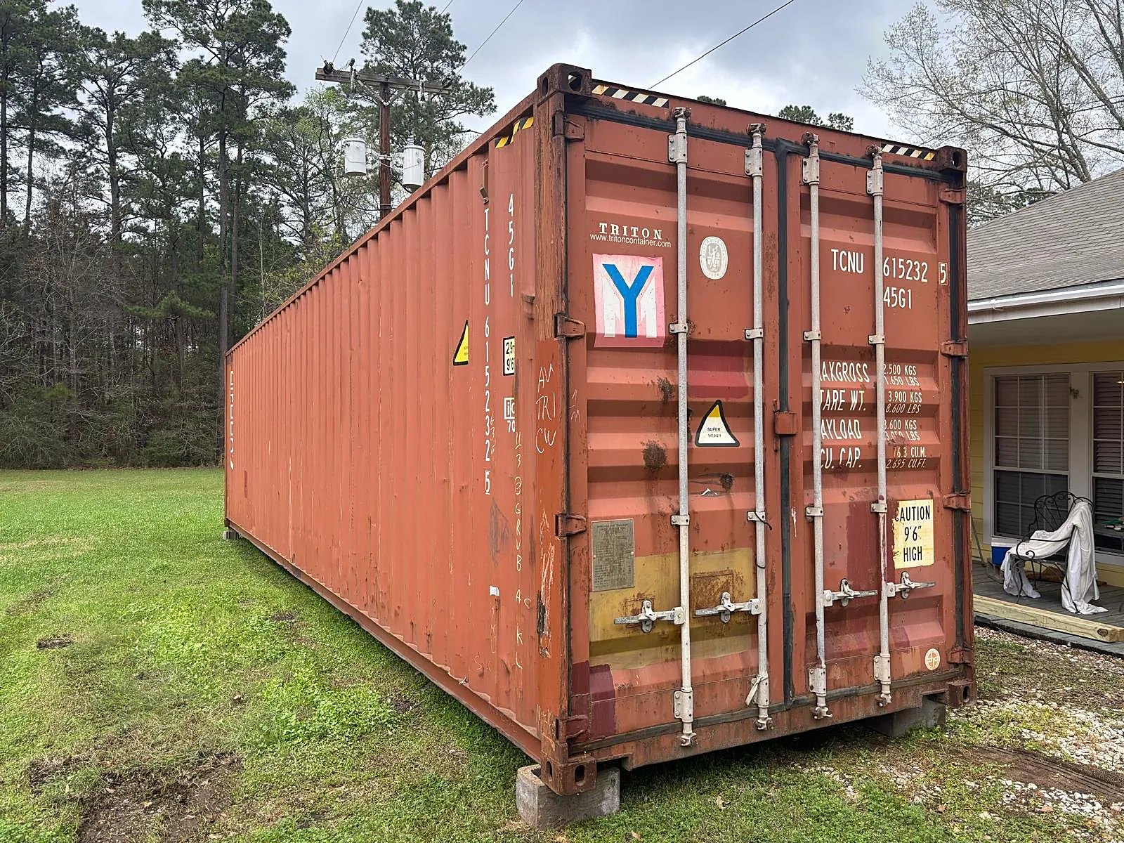 40ft High Cube Shipping Container – Used, Cargo Worthy – Weathered reddish shipping container with closed cargo doors on blocks beside a house