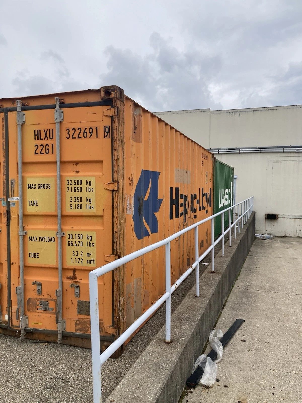 20ft Used Shipping Container – Ground-Level Storage – weathered orange shipping container beside railing and green container