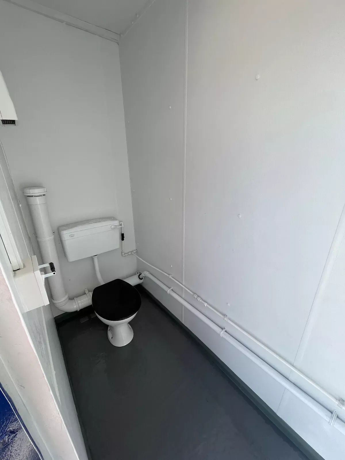 16ft × 9ft Modular Toilet Block – 3+1 Compartment Layout – narrow toilet cubicle with exposed plumbing and black toilet seat