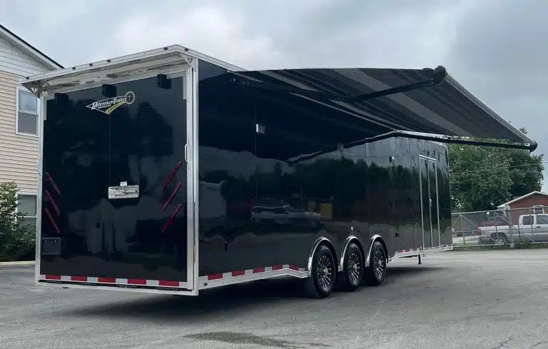 2025 Millennium 34 ft Car Trailer – Corner Bathroom – Black enclosed trailer rear and side view with a large striped electric awning extended