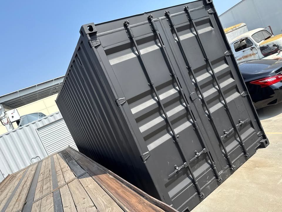 20ft Used Shipping Container – Matte Black, Side Roll-Up Door – black container with closed cargo doors beside parked vehicles