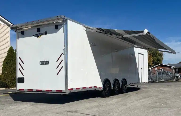 2025 Millennium 34 ft Car Hauler – Corner Bathroom – Rear three-quarter view of white trailer with extended striped awning and triple axles