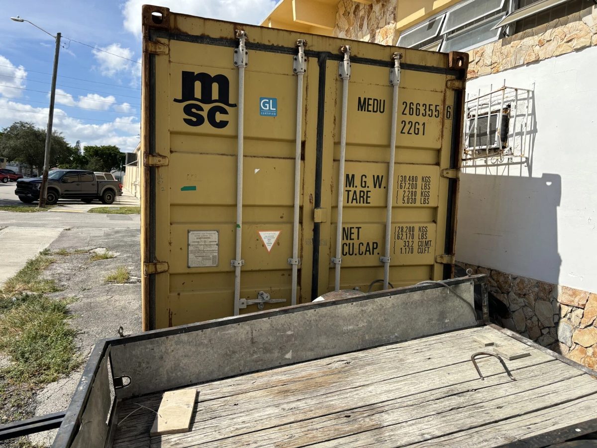 20ft Used Shipping Container – Yellow/Gold, Double Cargo Doors – Closed yellow container cargo doors beside a building, with a wooden trailer in front