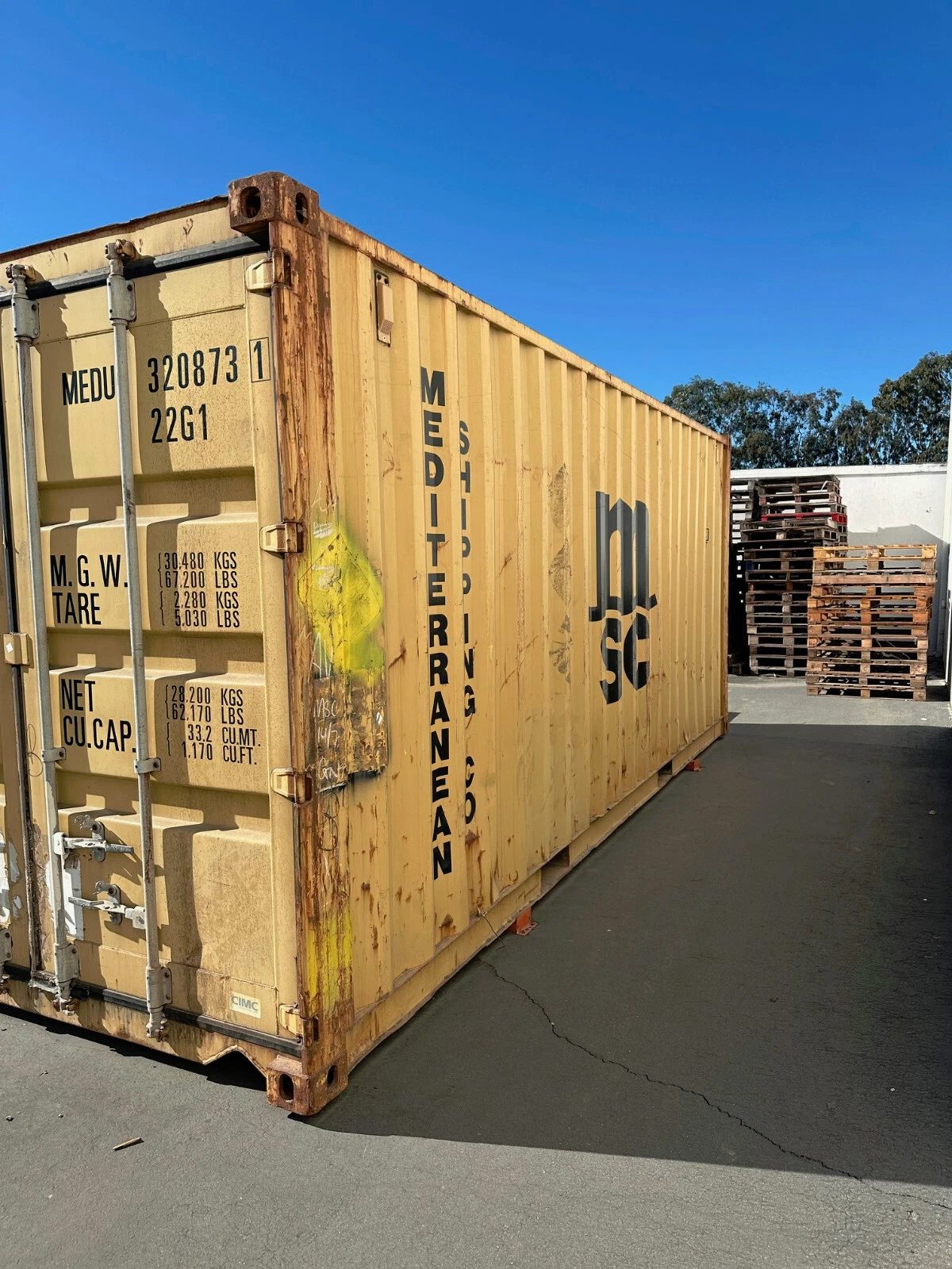 20ft Used Shipping Container – Yellow/Gold, Double Cargo Doors – Weathered yellow shipping container with closed cargo doors and rusted edges beside stacked pallets