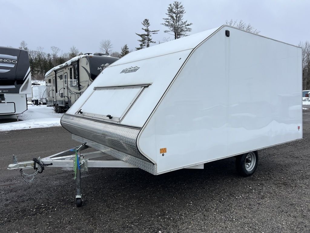 2022 High Country 12 ft Hybrid Trailer – Track Mats – A three-quarter exterior view of a white hybrid snowmobile trailer on a gravel lot