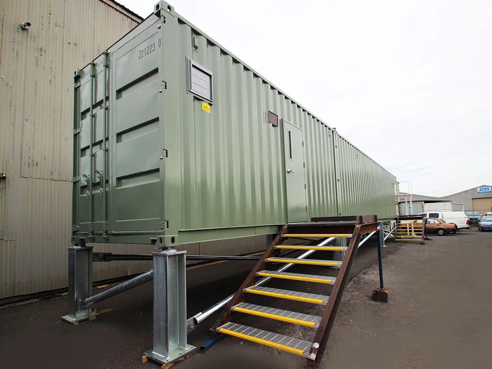 20 ft Switch Room Container – Insulated Panels – Long olive green container unit elevated on steel supports with an attached metal staircase