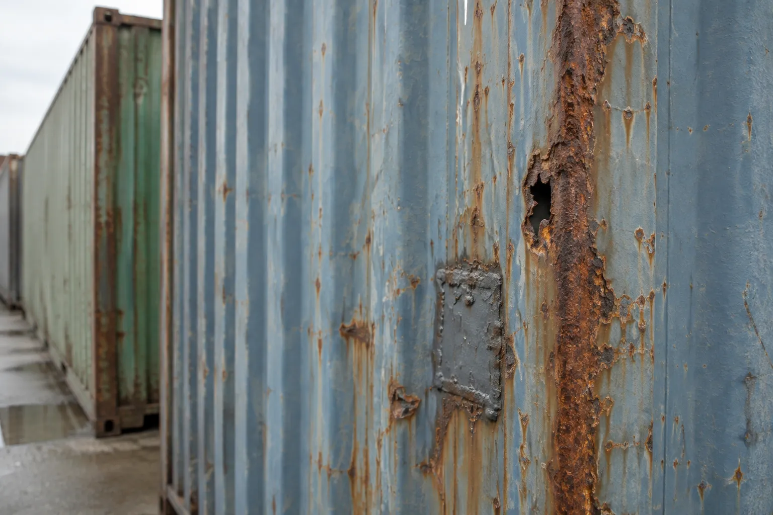 Used shipping container sidewall showing rust holes and an uneven welded repair patch.